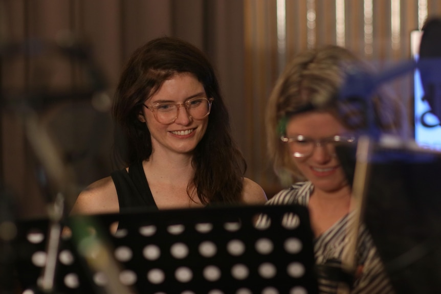 Kate McCartney and Kate McLennan wearing glasses and smiling, standing in front of music stands to record podcast Slushy