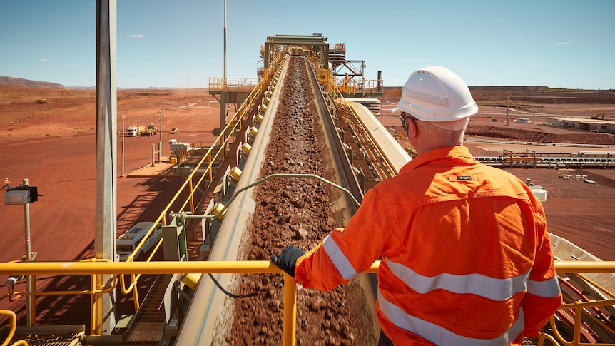 Man in high vis and hard hard looks over conveyor belt carrying iron ore at the South Flank mine.