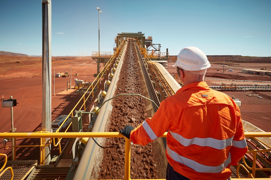 Un homme en haute visibilité et regardant durement la bande transporteuse transportant du minerai de fer à la mine South Flank.