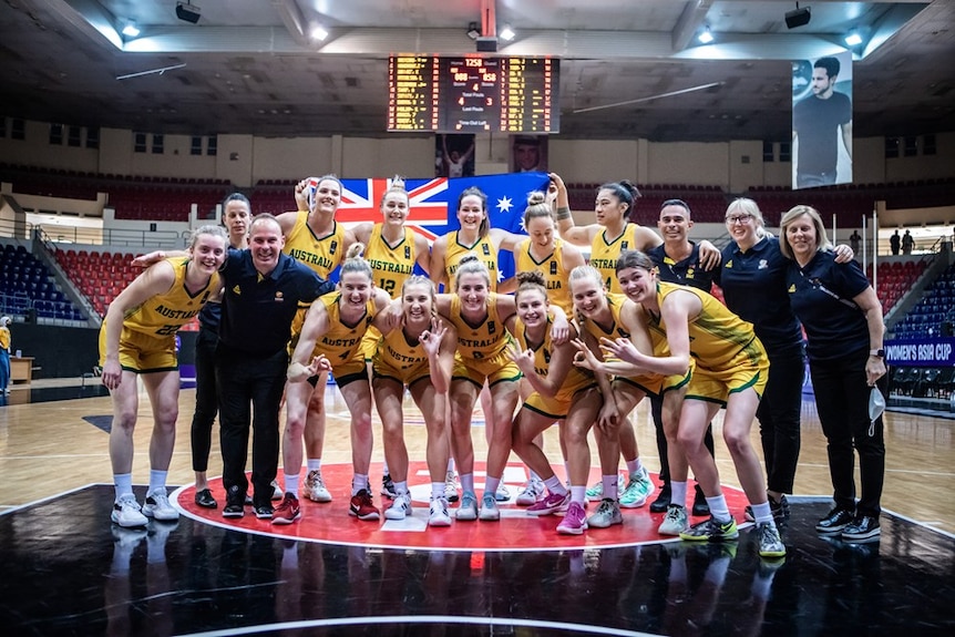 Members of the Australian women's basketball team pose for a photo, smiling, with the Australian flag behind them.