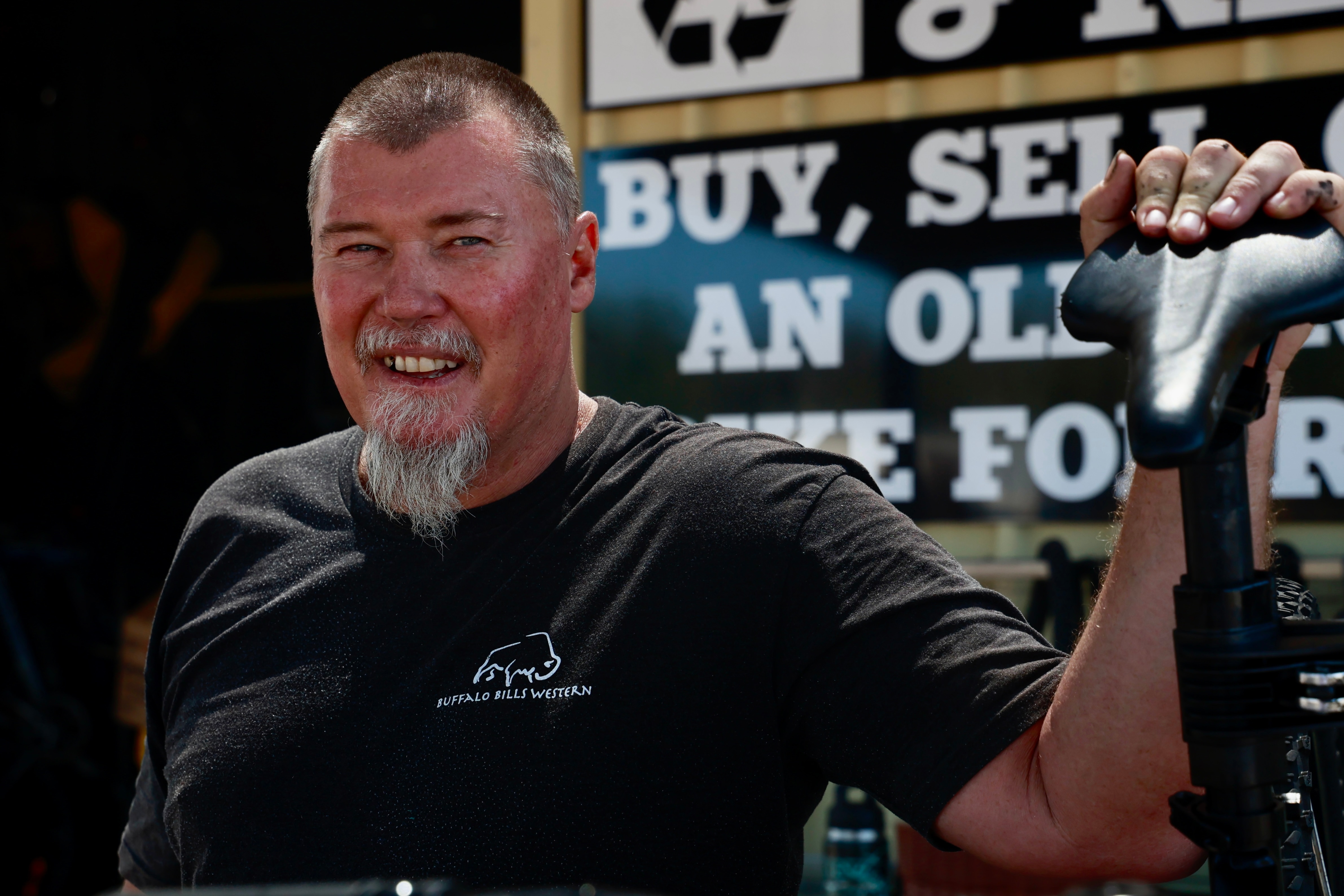 A smiling man faces holds a bike seat with shop signage behind him.
