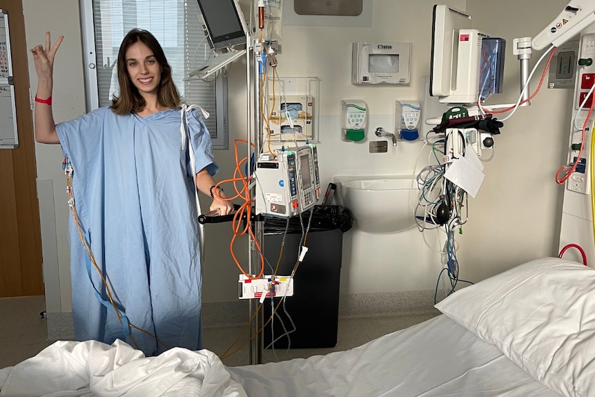 A young woman in a hospital gown standing next to a hospital bed, hooked up to machines and making a V sign