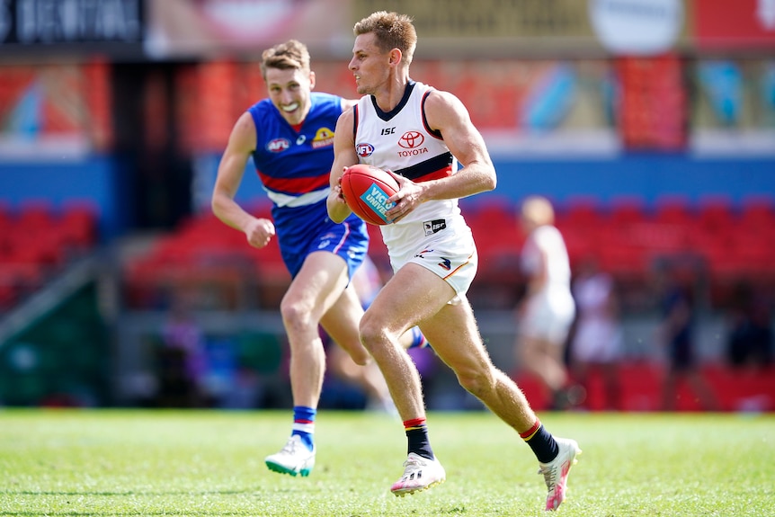An AFL player looks down the field, running with ball in hand as a defender trails behind him.