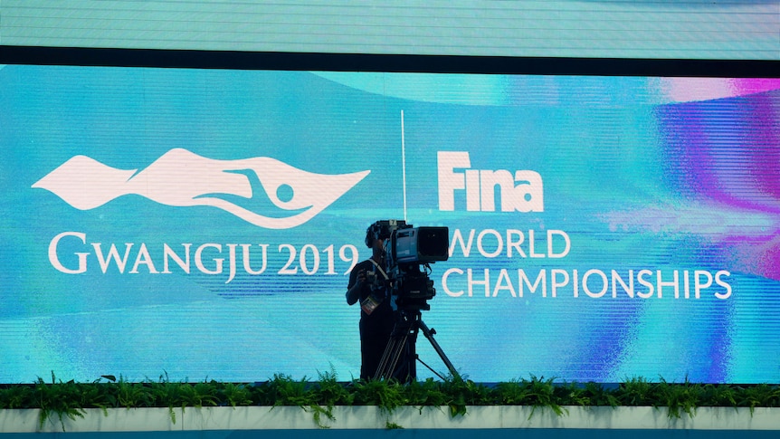 A TV cameraman stands in front of a big screen with a sign for FINA's world swimming championships.