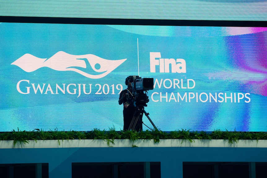A TV cameraman stands in front of a blue-coloured big screen with a logo and sign for the world swimming championships.