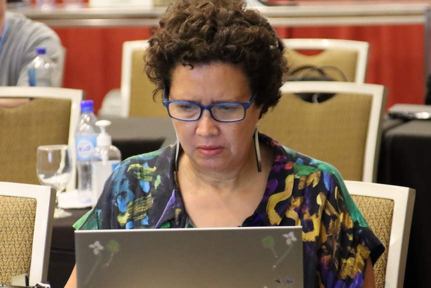 A fijian journalist with short curly brown hair and blue glasses sits at a desk on a laptop