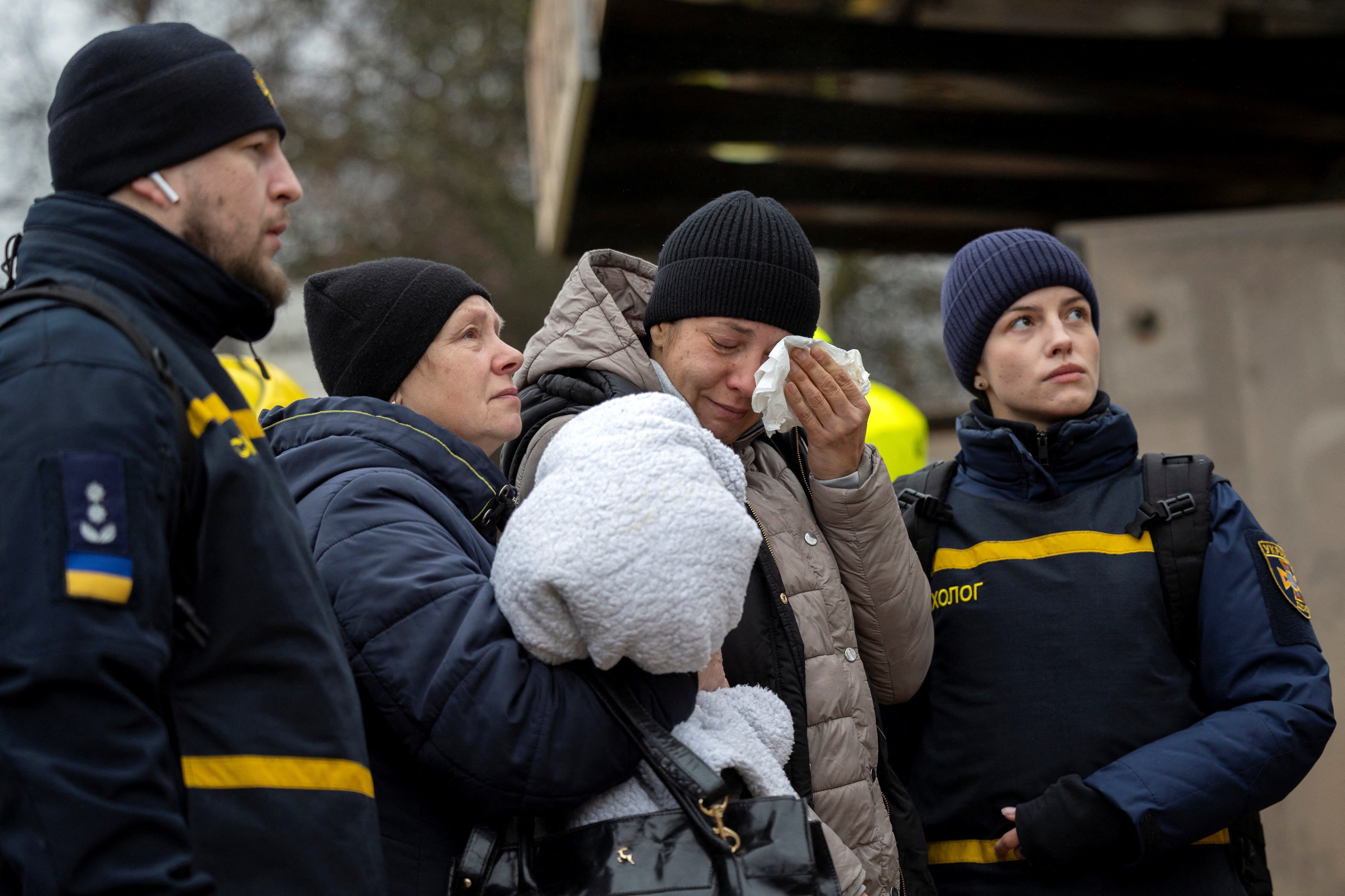 Two women in cold weather clothing look up with upset expressions as they are surrounded by Ukrainian firefighters.