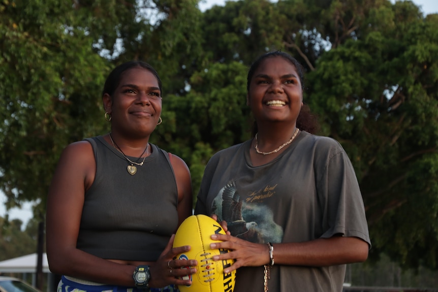 Whitney and Danielle Yunupiŋu hold a yelllow football and look across the camera while smiling against a backdrop of trees.