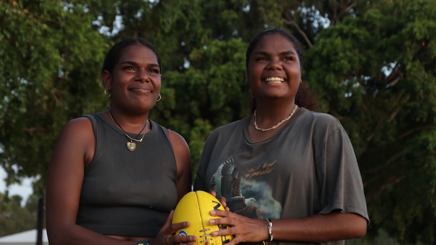 Whitney and Danielle Yunupiŋu hold a yelllow football and look across the camera while smiling against a backdrop of trees.