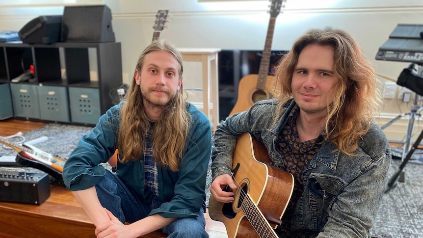 Two men with guitars sit on a stage casually