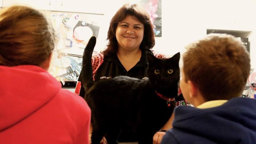 A woman stands facing with camera with a black cat and two children looking at her