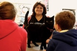 A woman stands facing with camera with a black cat and two children looking at her