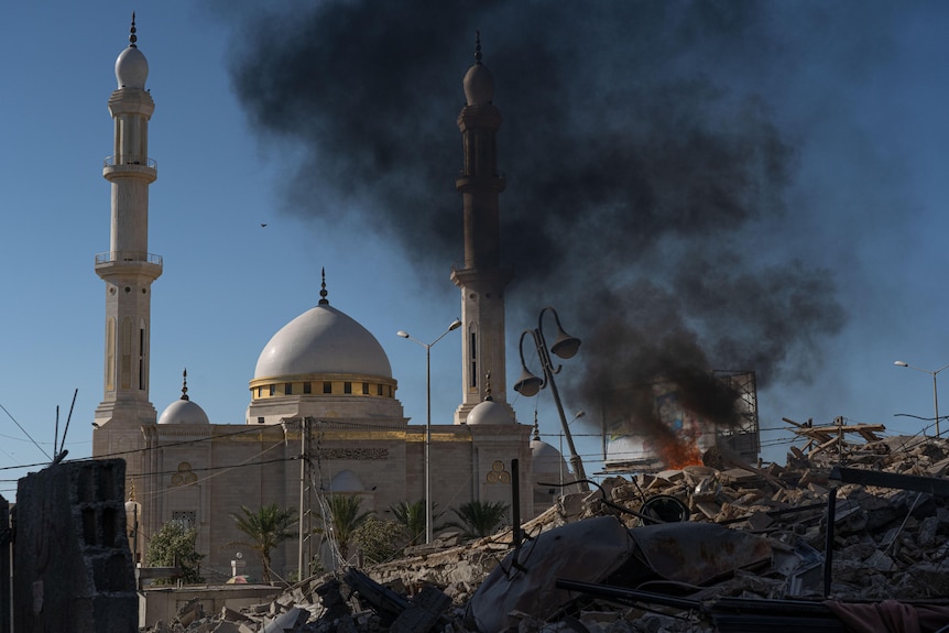 A mosque stands unscathed among rubble, with smoke drifting across the sky