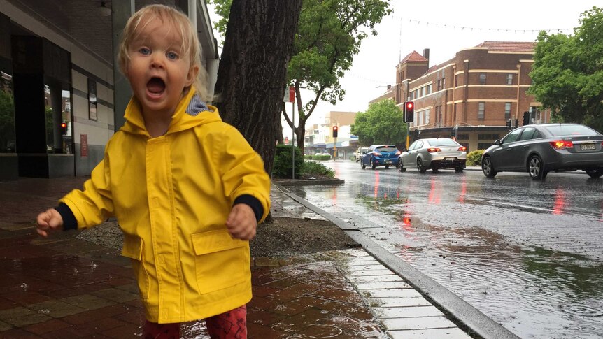 A young girl in a yellow raincoat jumps in a puddle on a street