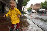 A young girl in a yellow raincoat jumps in a puddle on a street