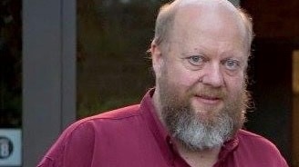 An older, bald man with a large red and white beard, wearing a dark red shirt.