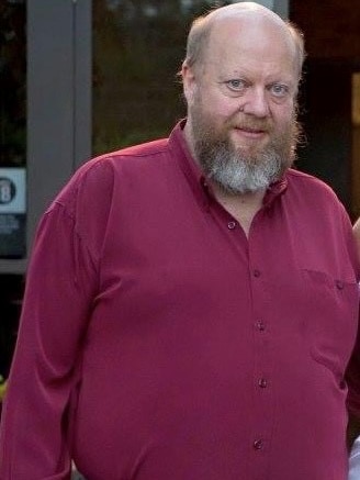 An older, bald man with a large red and white beard, wearing a dark red shirt.