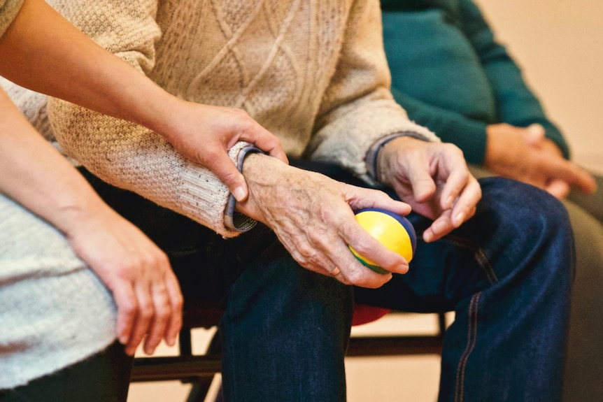 A young carer places her hand on the arm of a older man, holding a stress ball.