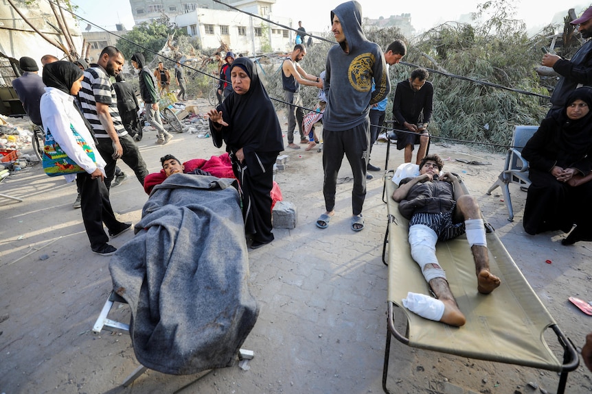 two young boys on stretchers outside with bandages and blankets on them and people standing around