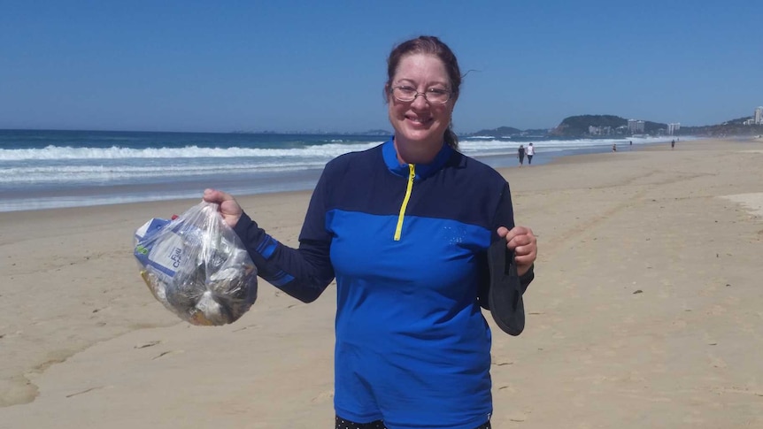 Woman stands on Gold Coast beach holding a bag of rubbish and a broken thong