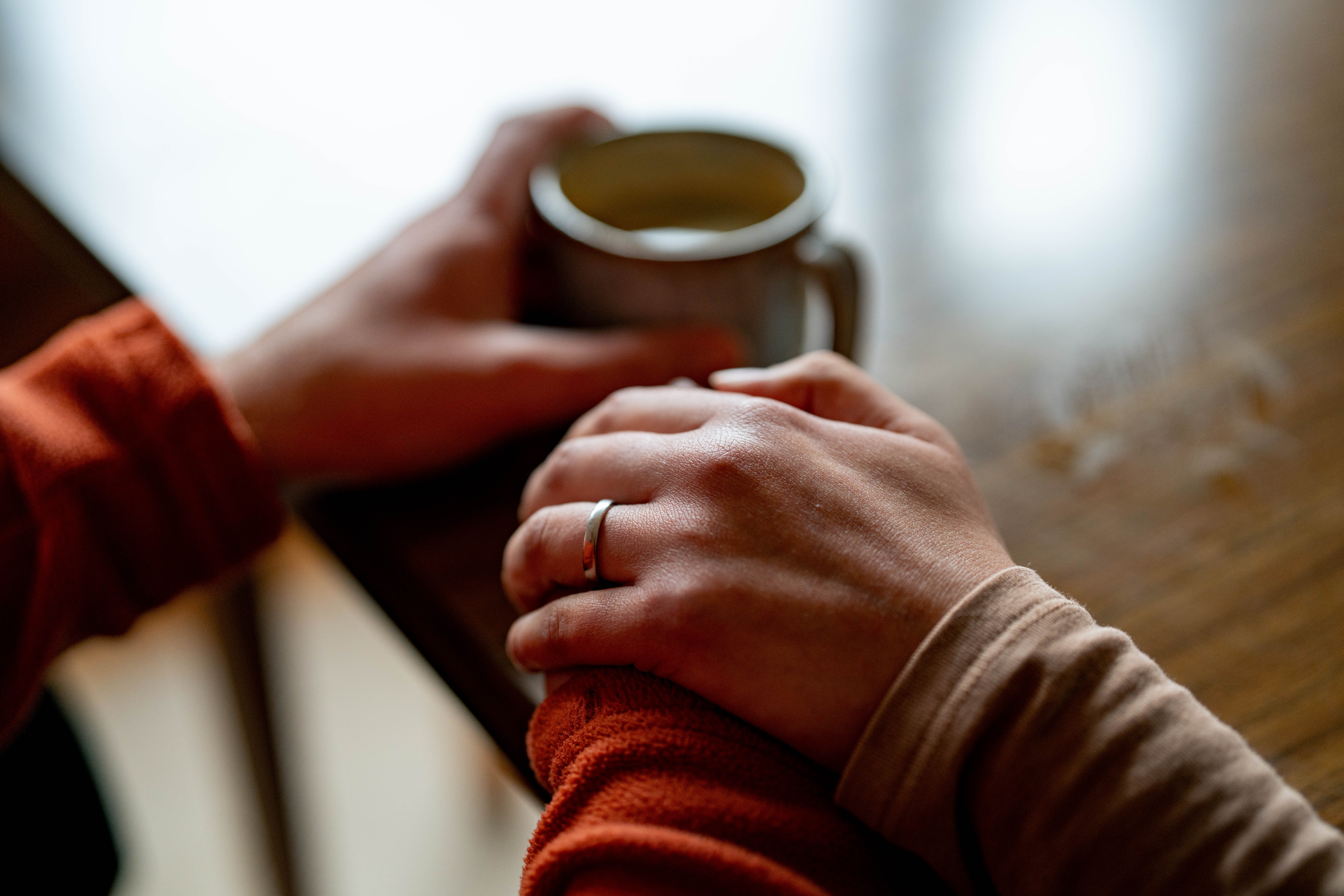 Close up of a woman's hand holding a man's hand, rested on a wooden dining table, the man holding a coffee in the other hand.