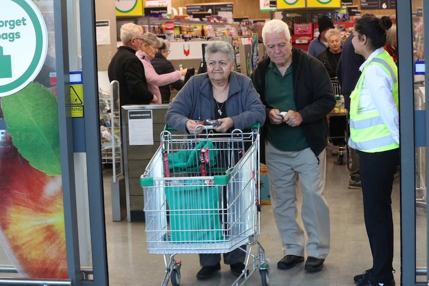 An older woman pushes a shopping trolley with a green bag inside it through a Woolworths. An older man walks next to her.