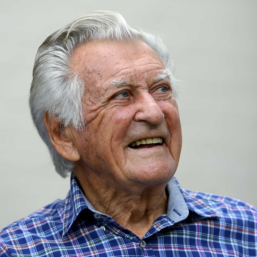 Bob Hawke looks to the right with a smile on his face. He wears a blue, purple and red check shirt and his hair is pushed back.