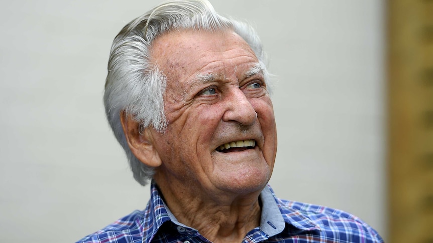 Bob Hawke looks to the right with a smile on his face. He wears a blue, purple and red check shirt and his hair is pushed back.