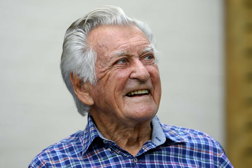 Bob Hawke looks to the right with a smile on his face. He wears a blue, purple and red check shirt and his hair is pushed back.