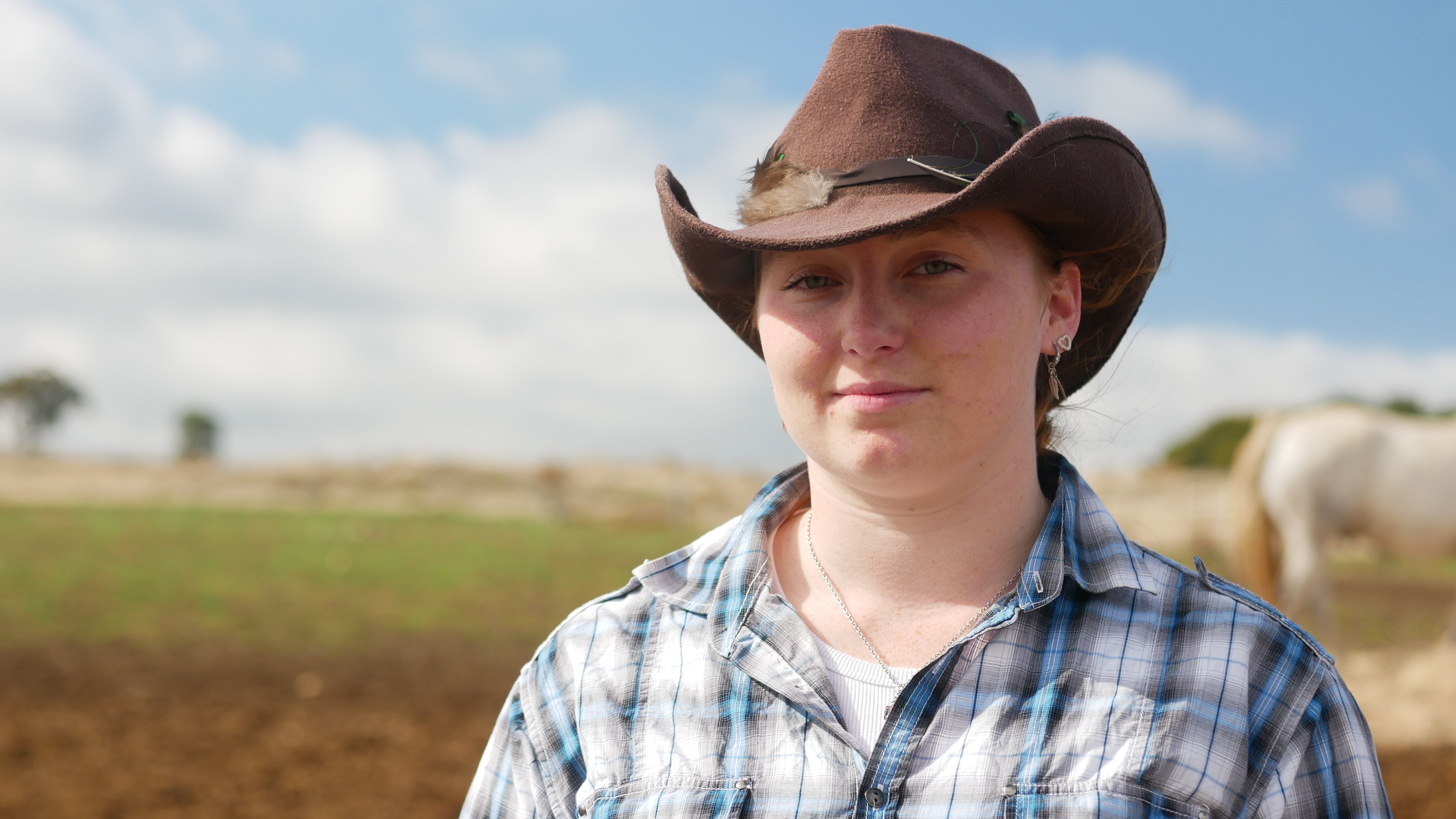 Young girl with an outback widebrim hat smiles into the camera. Theres is a white horse and a paddock in the background.