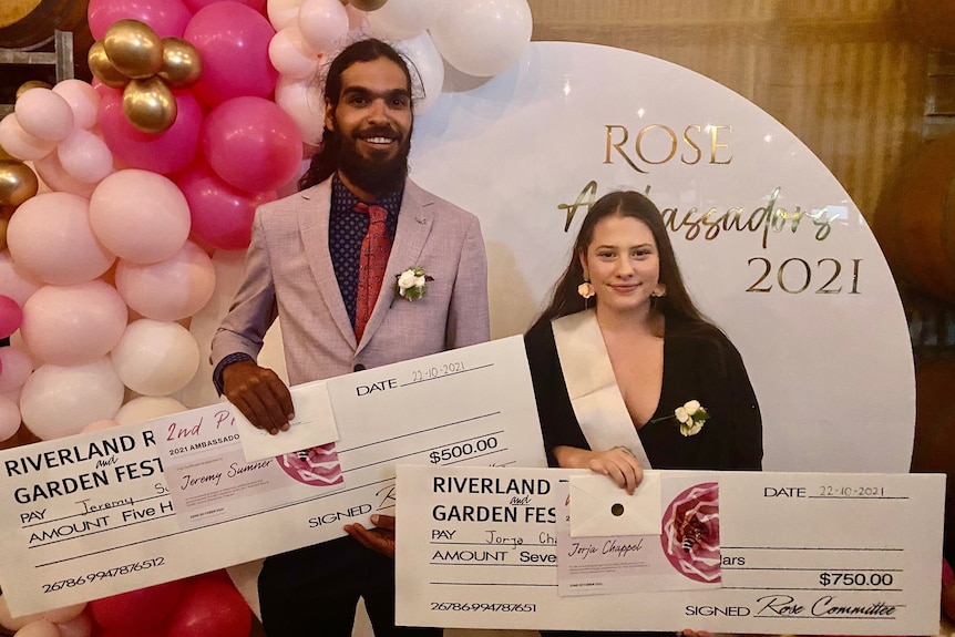 A man wearing a pink jacket and a woman wearing a black top and sash stand with giant cheques in front of pink balloons