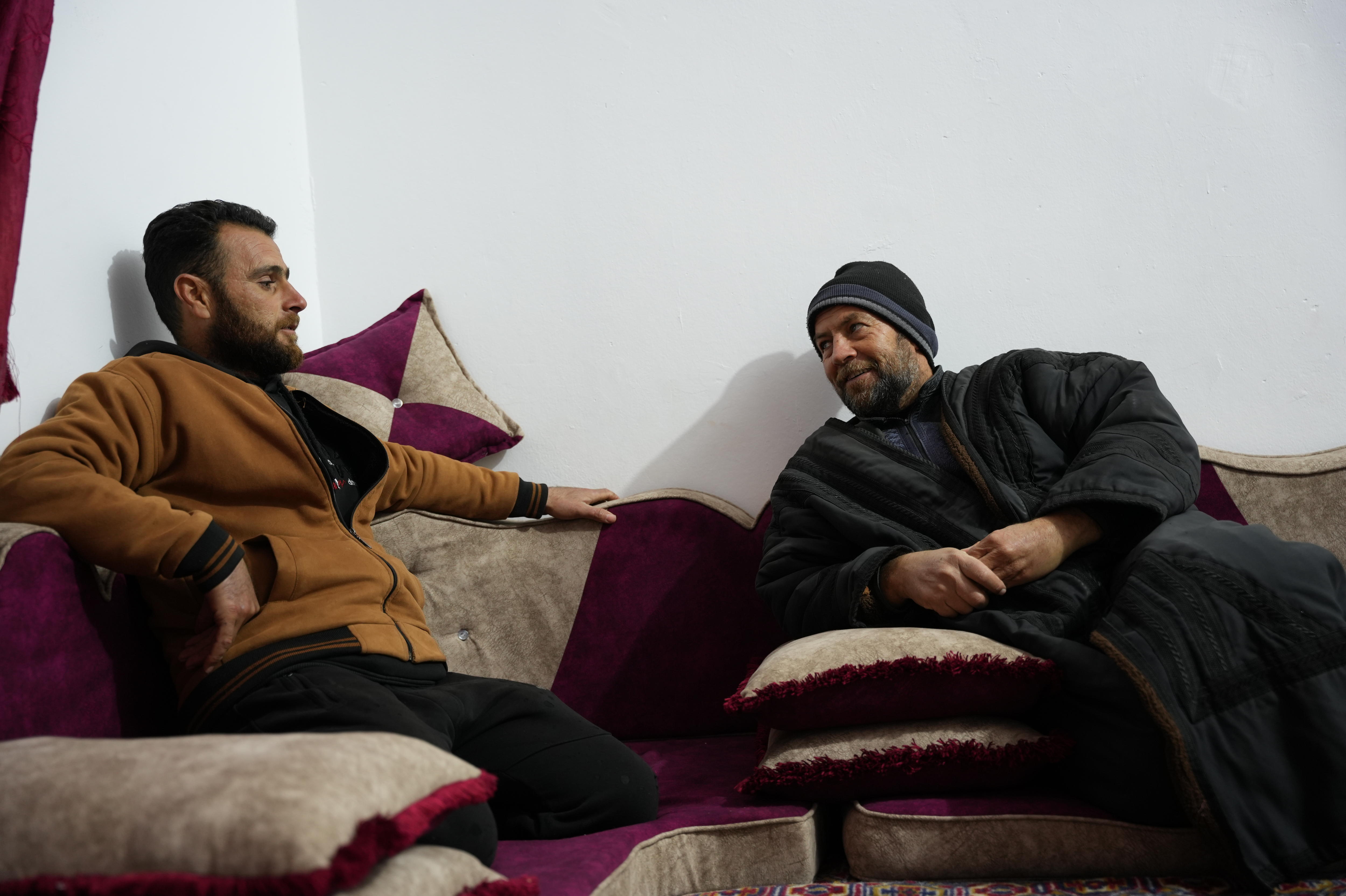 Dois homens sentados em um sofá, conversando e sorrindo.