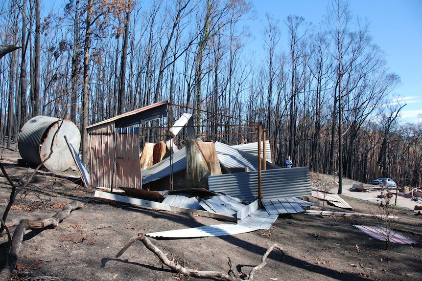 Sheets of metal lie on the ground next to a metal structure remains of a burnt out shed with charred forest in the background