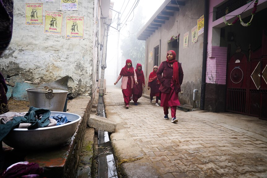 Gour women dressed in maroon walk down a cobblestoned street on a hazy day