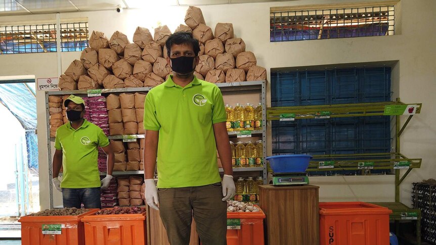 Two men in facemasks stand in front of food supplies packed in orange boxes