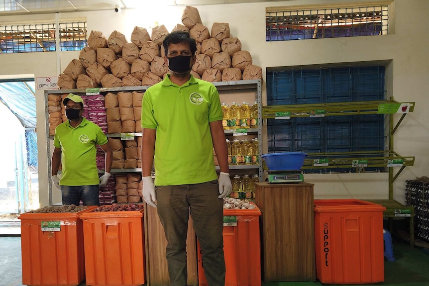 Two men in facemasks stand in front of food supplies packed in orange boxes