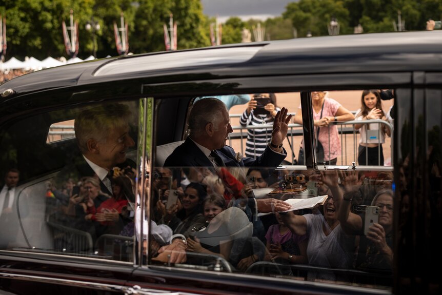 King Charles III waves out of a car window. A waving crowd is visible in the reflection.
