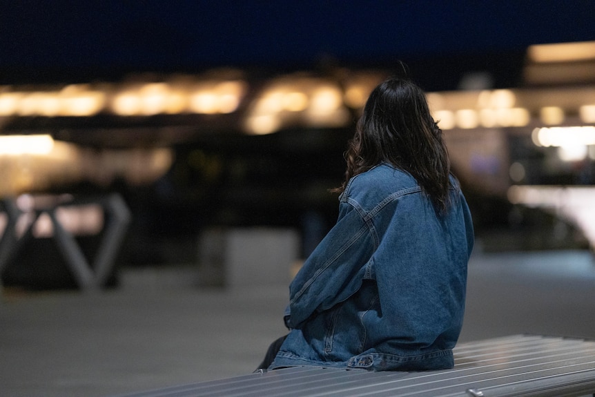 Woman in a denim jacket sitting at a wharf at night.