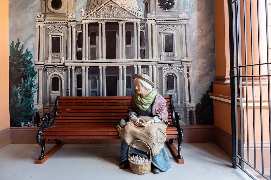 A life size figure of an elderly woman with a mural of a heritage building in the background.