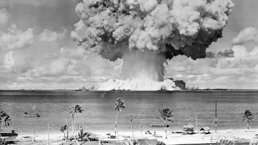black and white photo, mushroom shaped cloud above small island, palm-tree fringed coast in foreground