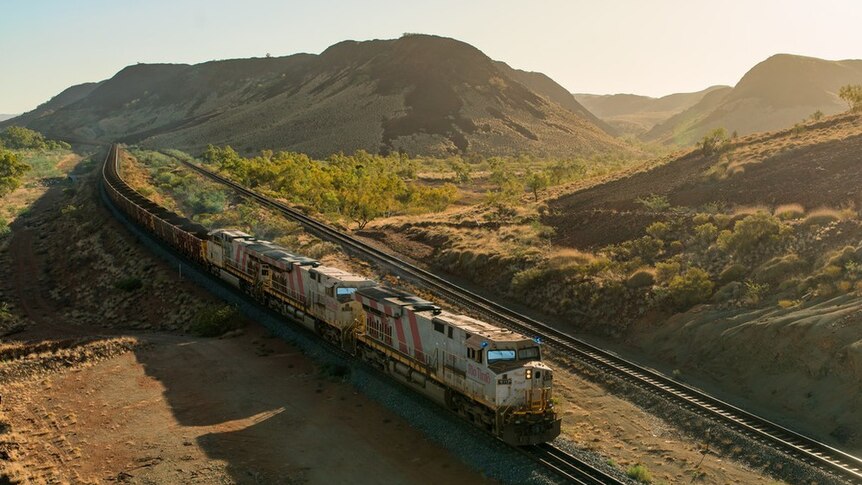 A train pulling dozens of carriages full of iron ore travels along a track in a valley of rolling red and green hills