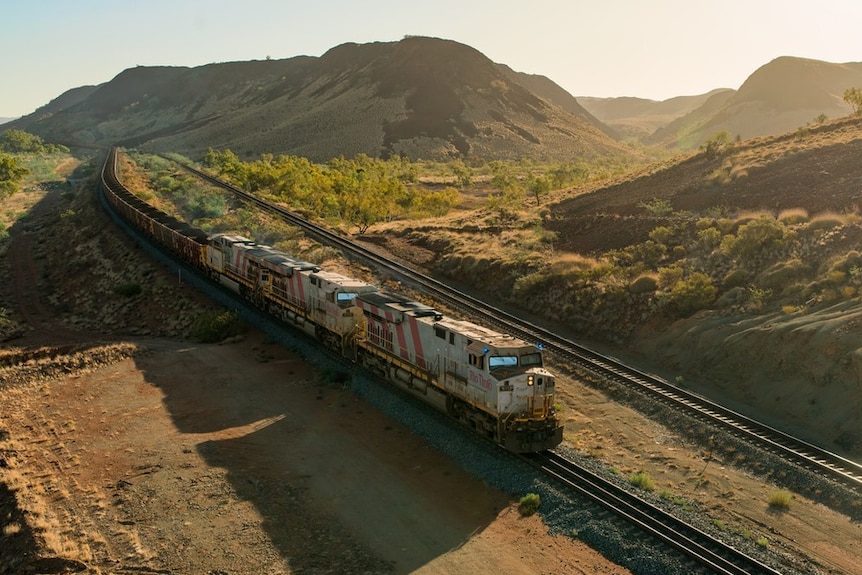 A train pulling dozens of carriages full of iron ore travels along a track in a valley of rolling red and green hills