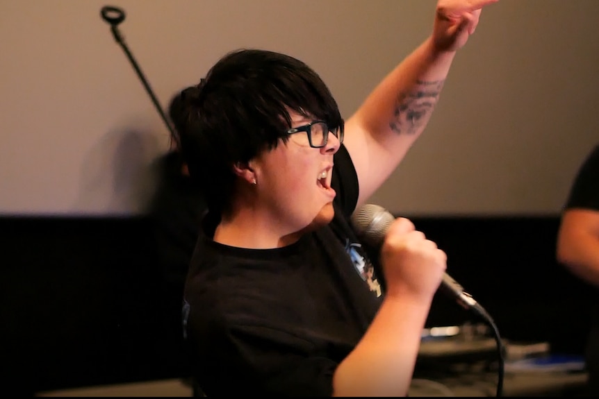 woman with short dark hair and an arm tattoo sings into a microphone