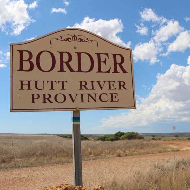 A white and red sign reading 'Border Hutt River Province' stands next to a gravel road under a blue cloudy sky.