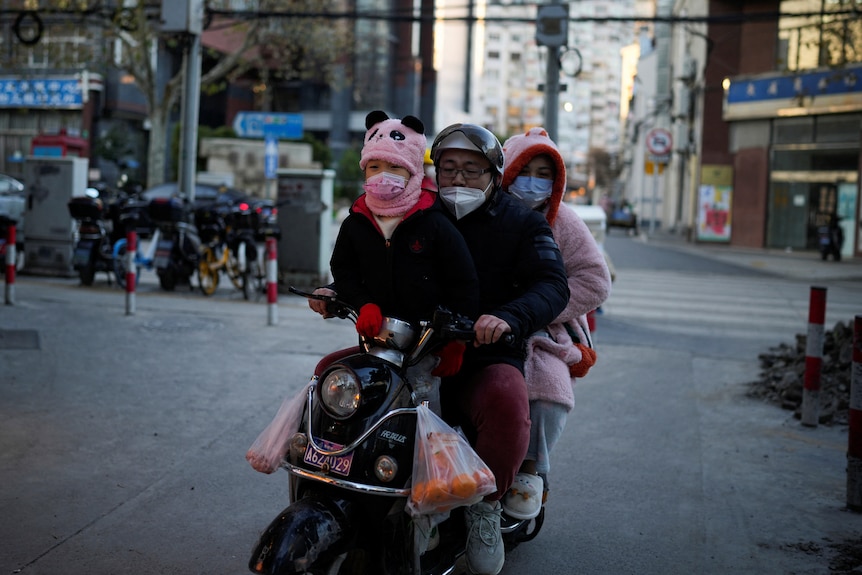 A family of three wear masks while riding a scooter through a street.