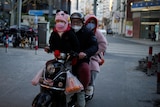 A family of three wear masks while riding a scooter through a street.