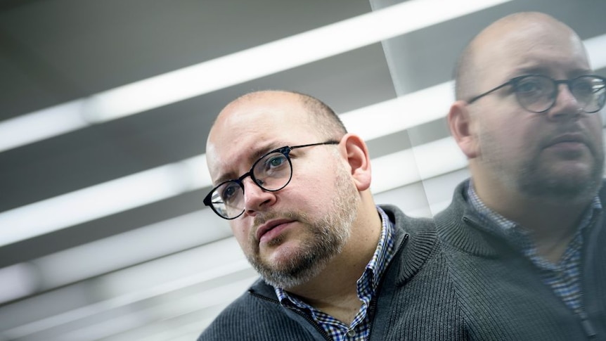 Jason Rezaian is pictured against ceiling lights above and a side mirror reflection of his face