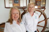 Two women stand in front of a big yacht wheel wearing white uniforms.