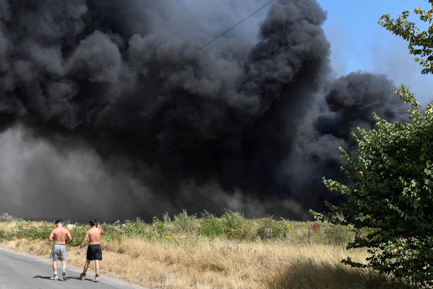 Two men walk down a road without shirts on. A big black plume of smoke fills the background. 