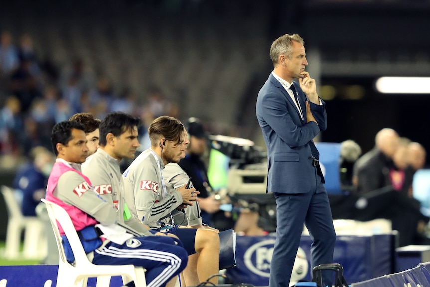 Grant Brebner stands with his hand to his chin as four men sit and watch behind him